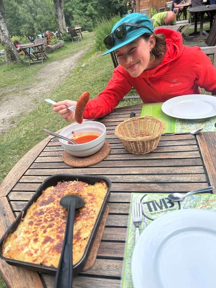 A female hiker dips a sausage into a bowl of sauce while sitting at an outdoor picnic table.